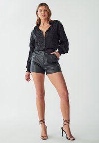 Zwarte kreukelige blouse met knoopsluiting en lange mouwen, gecombineerd met hoge zwarte leren shorts met zakken en stikdetails.