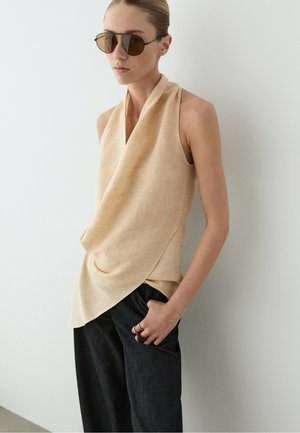 Frau trägt beige drapiertes ärmelloses Oberteil, dunkle Hose, Sonnenbrille und Ringe, mit der Hand in der Tasche, steht vor einer weißen Wand.