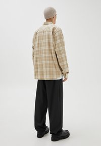 Persona con gorro de punto beige, camisa de cuadros beige oversize, pantalones negros de pierna ancha y zapatos negros, de pie de espaldas sobre fondo blanco.