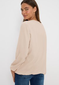 Beige, losvallende blouse van lichtgewicht stof, ronde halslijn, lange mouwen en subtiel textuurpatroon. Gedragen met blauwe denim jeans.