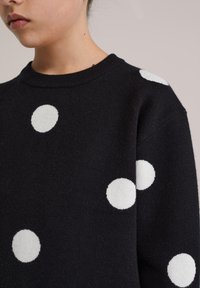 Pull noir à gros pois blancs, texture tricotée, encolure ronde côtelée, gros plan sur l'épaule et le haut de la manche.