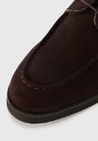 Dark brown suede shoe close-up, featuring a rounded toe, contrasting stitching, and black rubber sole. Smooth texture with a slight sheen.