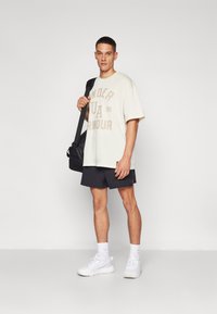 Camiseta de color beige claro con la marca "UNDER ARMOUR", combinada con pantalones cortos negros y zapatillas blancas. El sujeto sostiene un bolso negro de hombro.