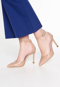 Nude lakované vysoké podpatky s špičatou špičkou, kotníkovým páskem a tenkým podpatkem. Na modrém pozadí.