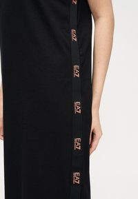 Κοντινό πλάνο ενός ατόμου που φοράει μαύρο φόρεμα με κάθετη μαύρη ρίγα που φέρει επαναλαμβανόμενα λογότυπα "EA7 Emporio Armani" σε ανοιχτό πορτοκαλί.