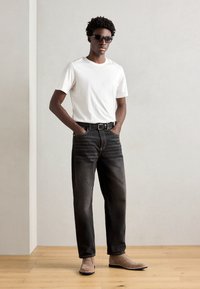 T-shirt bianco a maniche corte, jeans neri sbiaditi, cintura nera, stivali in suede marroni. Il modello indossa occhiali da sole scuri. Sfondo neutro.