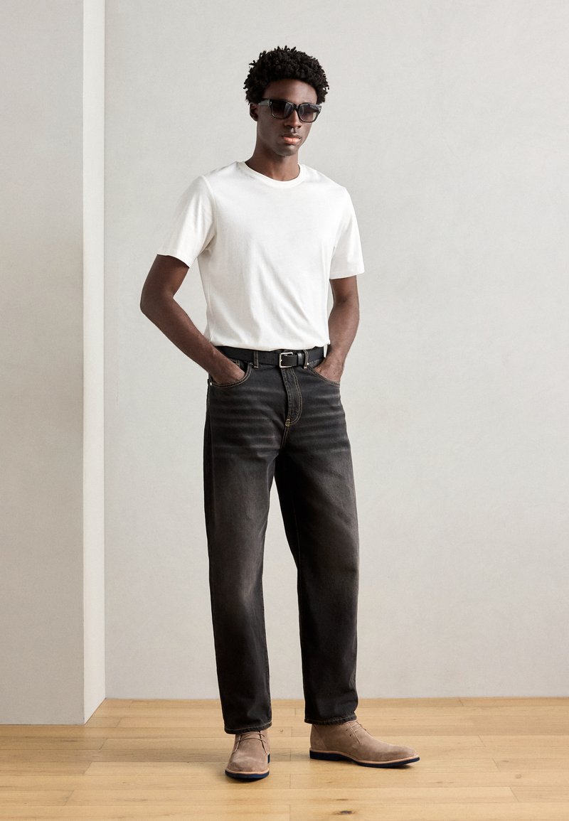 Weißes Kurzarm-T-Shirt, ausgebleichte schwarze Jeans, schwarzer Gürtel, braune Wildlederstiefel. Das Modell trägt dunkle Sonnenbrillen. Neutraler Hintergrund.