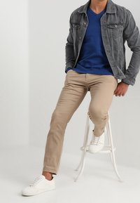 Grå denimjacka över en blå V-ringad tröja, beige byxor och vita sneakers. Modell sitter på en vit pall med en neutral bakgrund.