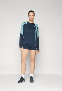 Marinfärgad träningsoutfit bestående av en kortärmad topp, shorts och en lätt jacka med turkosa detaljer och huva, tillsammans med svarta sneakers.