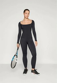 Γυναίκα σε μαύρο μπλουζάκι με μακριά μανίκια και κολάν, κρατώντας μία ρακέτα τένις, με φόντο ένα απλό ανοιχτό γκρι الخلفية.