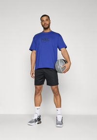 Mėlynas Nike marškinėliai su juodu logotipu, juodos šortai, balti kojinės ir raštuoti sportbačiai. Laiko krepšinį su tekstūruota paviršiumi.