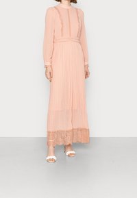 Robe longue plissée en tissu rose pâle avec des accents en dentelle et un col montant. Elle possède de longues manches et un délicat ourlet en dentelle. Portée avec des talons blancs.