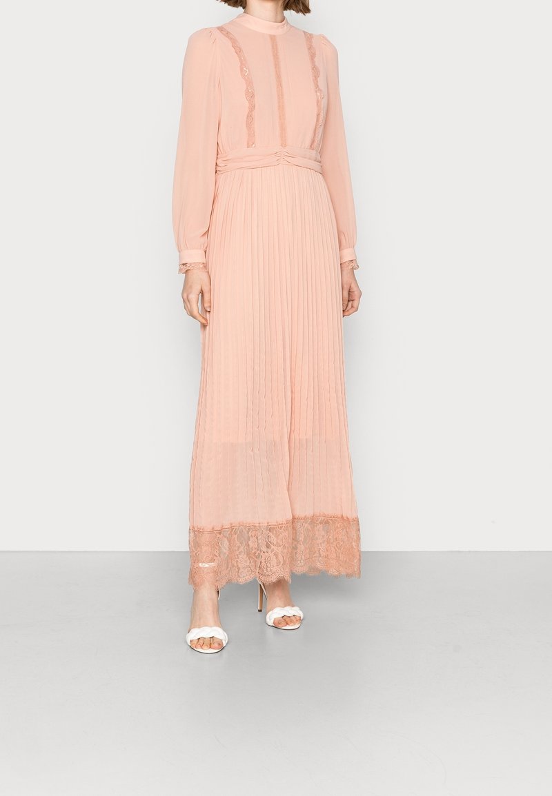 Robe longue plissée en tissu rose pâle avec des accents en dentelle et un col montant. Elle possède de longues manches et un délicat ourlet en dentelle. Portée avec des talons blancs.