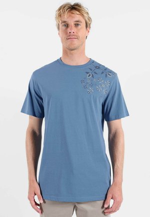 T-shirt en coton bleu à col rond, manches courtes, avec un motif floral dans des teintes plus foncées sur l'épaule droite. Texture lisse, coupe décontractée.