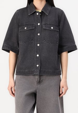 Person iført en sort, vasket denim kortærmet knap-op-skjorte med to frontlommer med klap og løse grå bukser.