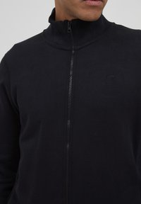 Svart zip-up jakke med høy krage, laget av et glatt stoff. Har en subtil logo og en teksturert glidelås. Tett passform.