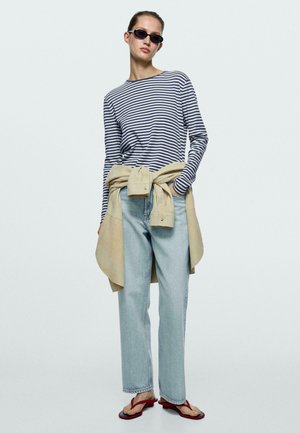 Frau mit schwarzen Sonnenbrillen, gestreiftem Langarmshirt, hellblauen Jeans, beiger Jacke, die um die Taille gebunden ist, und roten Sandaletten mit Absatz.