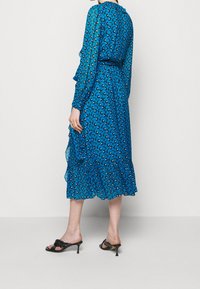 Vestido azul con estampado, hecho de tela ligera, con un dobladillo con volantes, mangas largas y cintura ceñida. Llevado con sandalias de tacón negras.