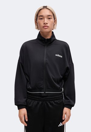 Person med kort blondt hår iført en sort Adidas træningsjakke og matchende sorte Adidas bukser mod en ensfarvet lys baggrund.