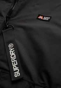 Zwarte zip-up jas met een gestructureerd oppervlak, voorzien van een prominent Superdry-logo en een gemarkeerde band. Rits- en stikdetails zichtbaar.