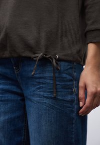 Maglietta grigia a righe con coulisse in fondo e dettagli in argento, abbinata a jeans di denim blu con cuciture laterali e una texture liscia.