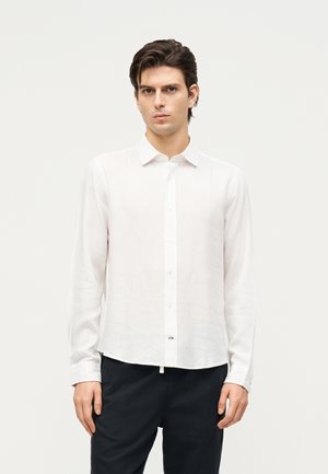 Noor mees, kellel on seljas valge pika varrukaga nööbitav särk ja mustad püksid, seisab ühetaustalise heleda tausta ees.