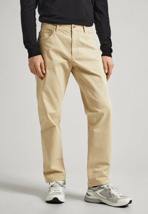 Pantalones de algodón beige con un diseño de pierna recta, estilo de cinco bolsillos y una clásica cremallera y botón. Combinados con zapatillas deportivas grises.