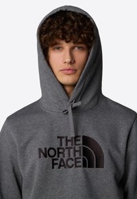 Sudadera gris con capucha ajustable, que presenta un gráfico de logo oscuro en el pecho. La tela parece suave con un acabado texturizado.