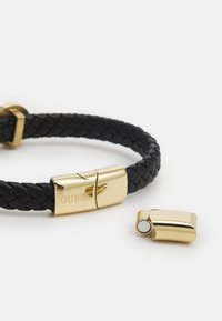 Black braided leather bracelet featuring a gold clasp with "GUESS" engraving and a detachable gold accent piece.