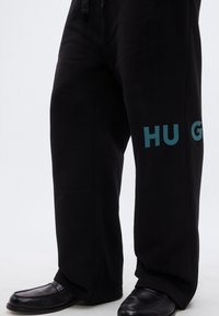 Μαύρο φούτερ παντελόνι με χαλαρή εφαρμογή, με τοιρκονέ μπλε λογότυπο "HUG" στο αριστερό πόδι. Το ύφασμα φαίνεται μαλακό με ομαλή υφή.