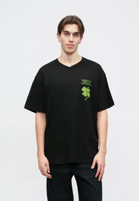 Czarna bawełniana koszulka t-shirt o dużym kroju, z zielonym haftowanym czterolistnym koniczyną i napisem na górnej lewej stronie.