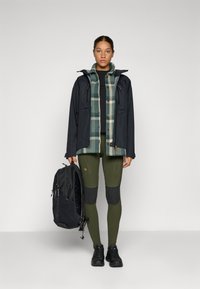 Svart vattentät jacka med huva, grön rutig skjorta, svart topp, olivgröna leggings med knälappar, svarta sneakers och en svart ryggsäck.
