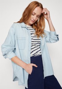 Camicia di jeans azzurro chiaro con vestibilità rilassata, maniche lunghe e una tasca sul petto, sovrapposta a una maglietta a righe. Pantaloni blu navy.