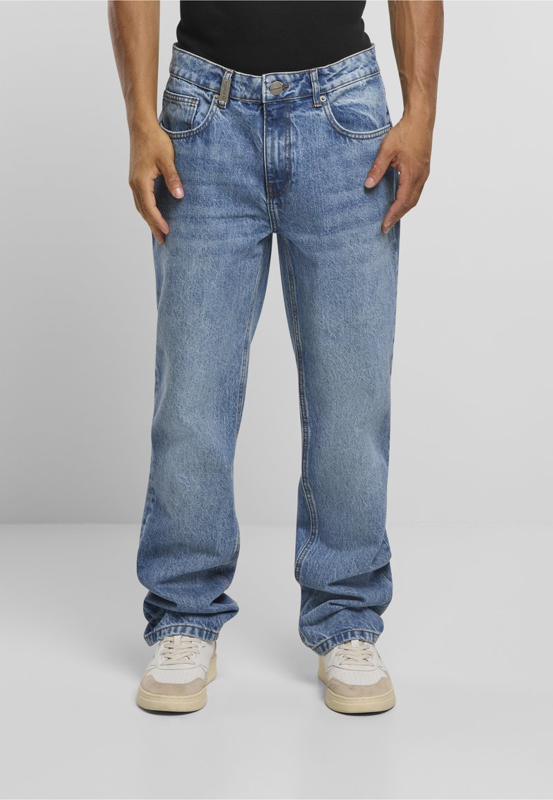 Személy laza, világoskék farmernadrágban és fehér sportcipőben, egy egyszínű világosszürke háttér előtt áll.