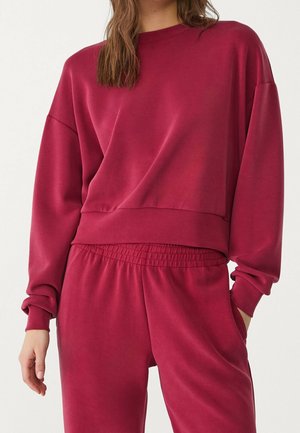 Personne portant un sweat-shirt ample rouge foncé et un pantalon assorti avec une taille élastique, une main dans la poche sur fond blanc.