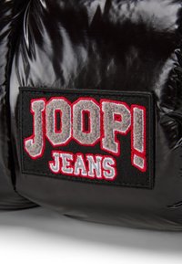 Črna, sijoča puffer jakna z teksturiranim črnim žepom, na katerem je izvezena oznaka "JOOP!" v sivi in rdeči barvi ter "JEANS" v beli barvi.