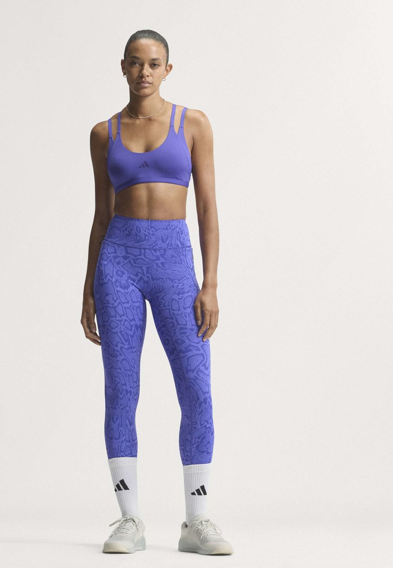 Femme debout en leggings bleus à motifs, brassière de sport bleue assortie, chaussettes blanches et baskets blanches, sur un fond uni.