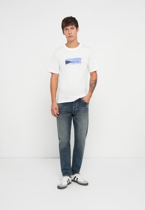 Young man standing, wearing a white graphic t-shirt, blue jeans, white sneakers, with a tattoo on his left forearm.