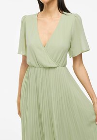 Femme portant une robe plissée vert clair à manches courtes et décolleté en V, tenant légèrement la jupe sur le côté.