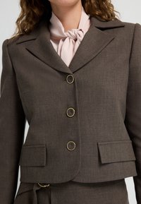 Brązowy żakiet z klapami, trzema złotymi guzikami i kieszeniami z przodu. Noszony na jasnoróżowej bluzce z zawiązanym krawatem. Gładka tekstura.