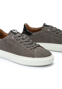 Zapatillas de ante gris con suelas de goma blanca, cordones grises y un acento en el talón de cuero negro. Presentan un diseño minimalista y detalles del logotipo.