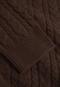 Maglione marrone lavorato a maglia con motivo a treccia e polsini a coste, realizzato in un materiale morbido con una texture pelosa. Dettaglio in primo piano mostrato.