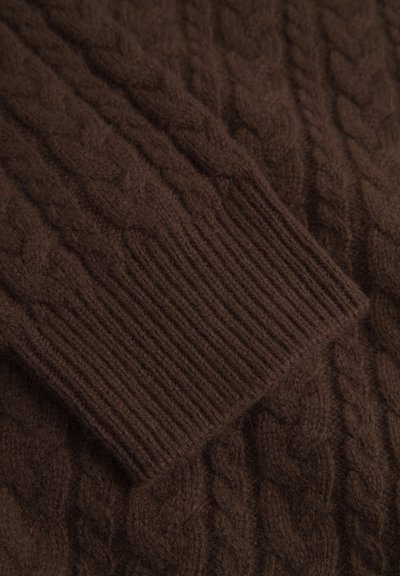 Rjav pleten pulover z vzorcem kablov in rebrastimi manšetami, narejen iz mehkega materiala z kosmatim otipom. Prikazan je bližnji posnetek detajla.
