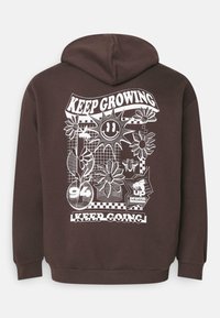 Brun hette med et stort hvitt grafikk på baksiden, som viser blomster, en smilende sol, og tekstdesignene "KEEP GROWING" og "KEEP GOING."