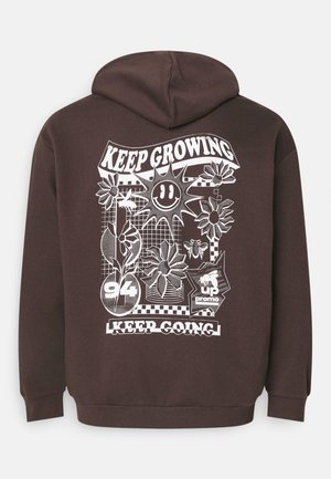 Brun hette med et stort hvitt grafikk på baksiden, som viser blomster, en smilende sol, og tekstdesignene "KEEP GROWING" og "KEEP GOING."