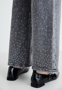 Pantalones de mezclilla gris de pierna ancha adornados con pequeñas embellishments en forma de perlas, espaciadas de manera uniforme. Combinados con zapatos planos negros brillantes.