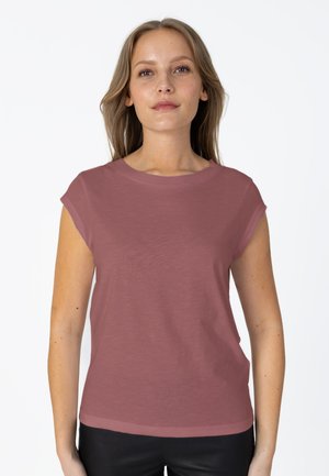 Jeune femme aux longs cheveux châtain clair porte un t-shirt rose poudré à manches courtes, debout devant un fond blanc uni.