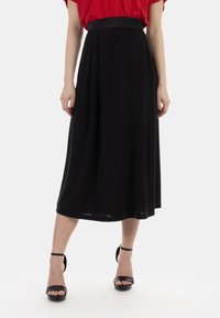 Gonna nera midi con un elastico lucido in vita, caratterizzata da una texture liscia e una silhouette fluida. Abbinata a décolleté nere con cinturino alla caviglia.