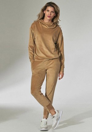 Beżowy dres z weluru, składający się z bluzy z kapturem i kołnierzem oraz zwężanych spodni. Uzupełniony białymi sneakersami. Gładka faktura, minimalne akcenty.