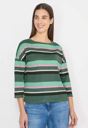 Vrouw draagt een lange mouw shirt met groene, roze, zwarte en witte strepen en een blauwe spijkerbroek, glimlacht en kijkt opzij.